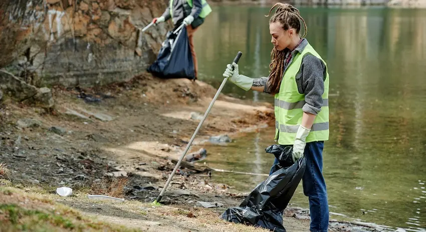 Two volunteers in safety vests use litter pickers to collect trash along a polluted riverbank representing community service work commonly assigned as a condition of probation or deferred adjudication in lieu of jail time in Texas.