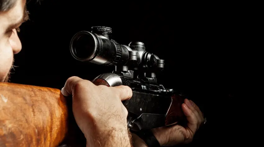 A bearded man aims a scoped bolt-action rifle in a darkened setting depicting the type of firearm use and possession that can lead to an investigation and federal gun charges depending on the circumstances surrounding the weapon in The Woodlands.