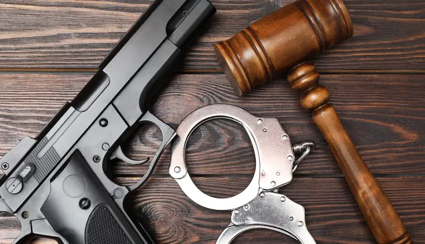 A black semi-automatic handgun- a pair of steel handcuffs- and a wooden judge's gavel arranged together on a dark wood grain surface- representing the intersection of criminal law enforcement and the judicial process in cases involving federal gun charges in The Woodlands with help from Cowboy Law Group.
