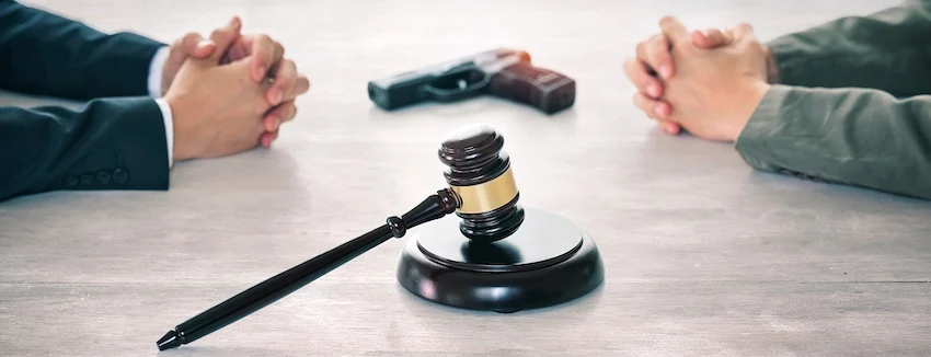 A client and an attorney sit across from each other at a table with a handgun and a judge's gavel in the foreground representing federal cases involving gun manufacturers and firearms-related legal proceedings at Cowboy Law Group.