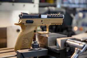 A tan and black semi-automatic pistol rests on a metal machining vise inside a manufacturing facility showcasing the precision equipment gun manufacturers use during the production process in The Woodlands TX.