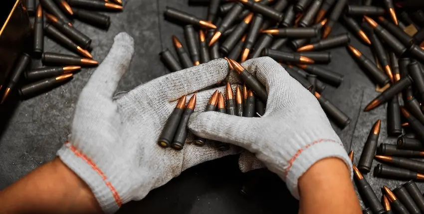 A worker wearing gray cotton gloves sorts and handles multiple rifle cartridges spread across a dark surface reflecting the scale of ammunition production managed by licensed gun manufacturers in The Woodlands TX.